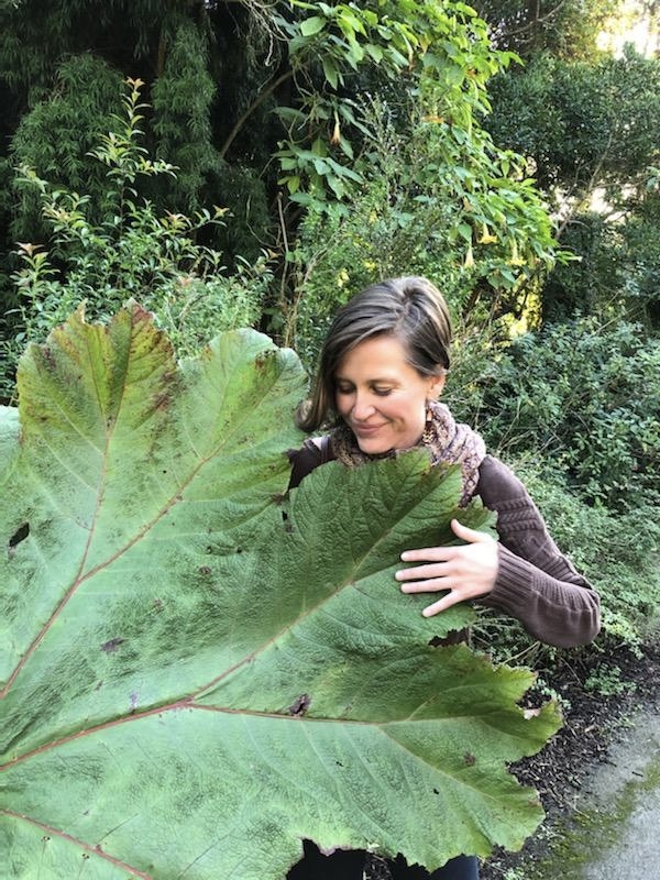 Jen Poole, founder of Sacred Bridge, smiling and embracing a giant leaf in a lush garden