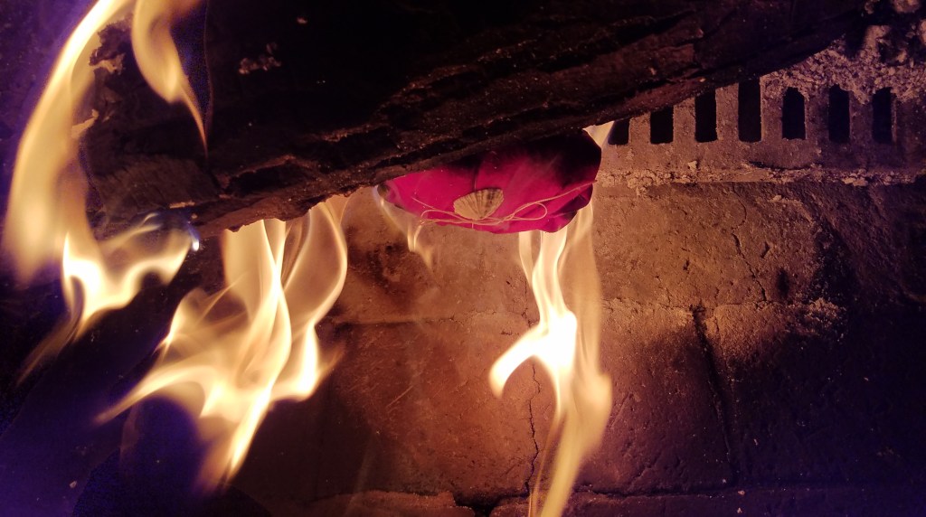 A pink cloth offering bundle tied with string and a shell, resting on a burning log in a fireplace