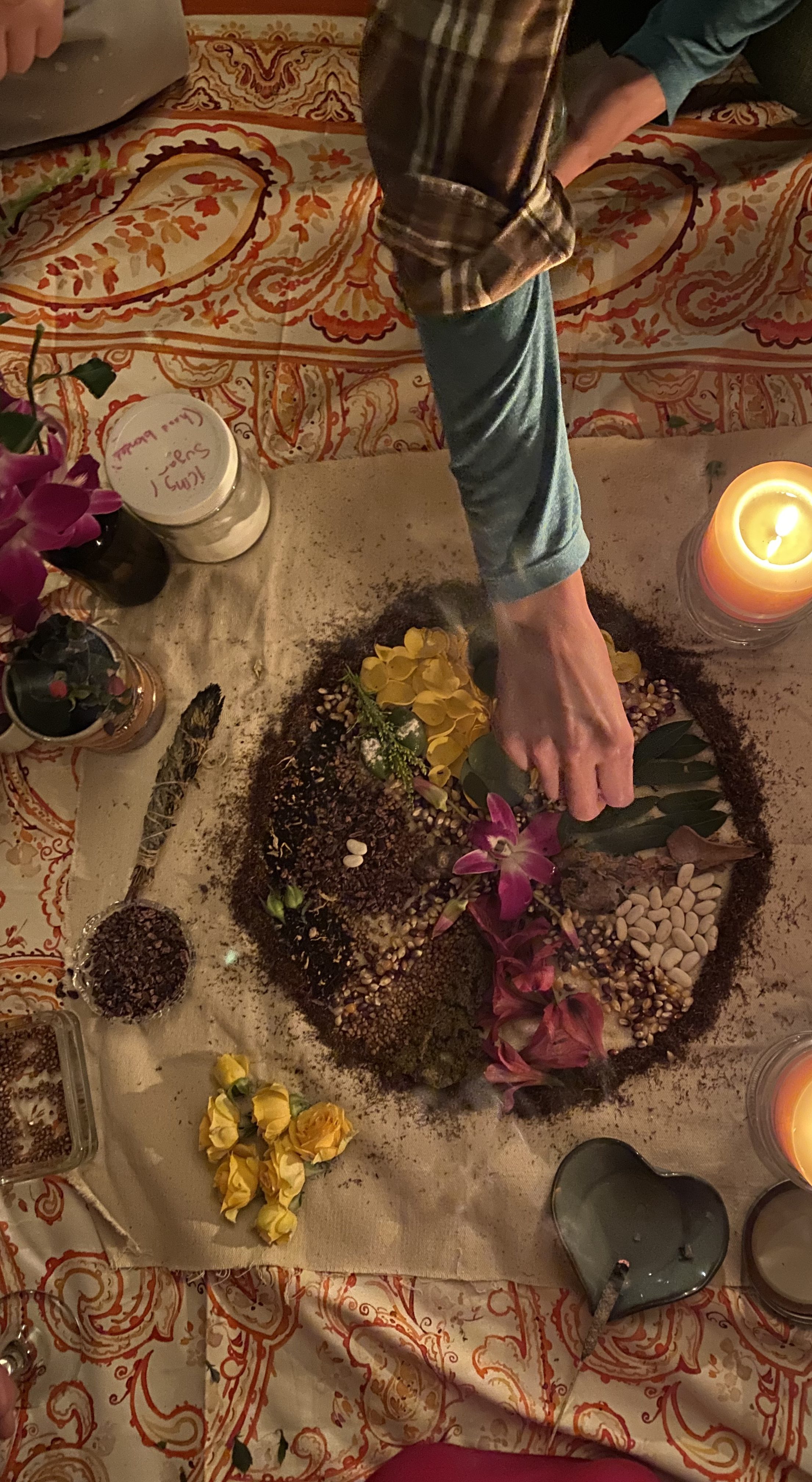 Hands placing flowers and herbs into a circular offering on a patterned cloth, surrounded by candles