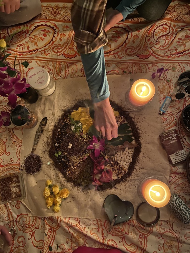 Hands placing flowers and herbs into a circular offering on a patterned cloth, surrounded by candles